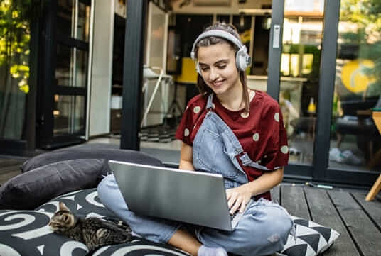A woman sits outdoors on her deck and uses her laptop in her lap.