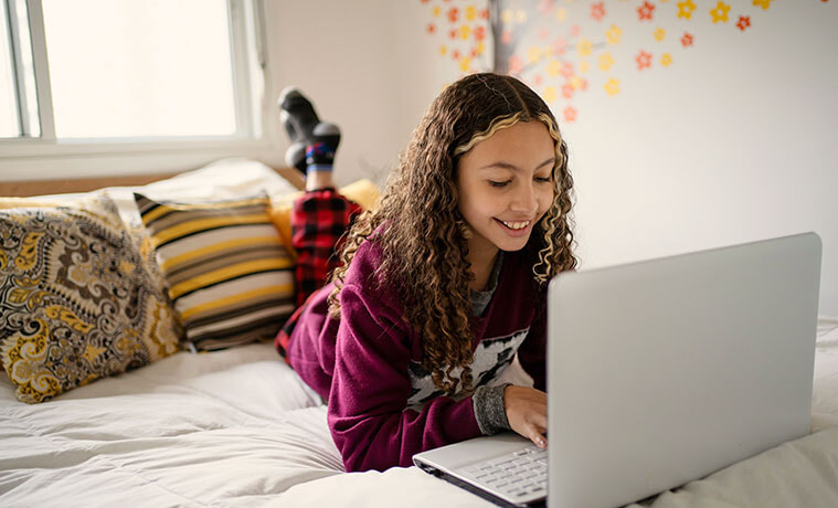 A teenage girl lie on her bed while using her laptop.
