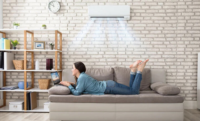 A woman lays on her sofa and uses her laptop while her split system air conditioner keeps her cool.