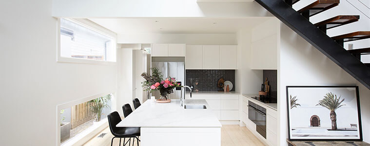 Side-view of light-flooded white kitchen with black stools and black staircase.